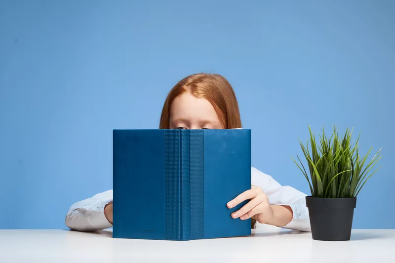 Рыжеволосая девушка сидит за столом с книгой в руках и учит уроки
