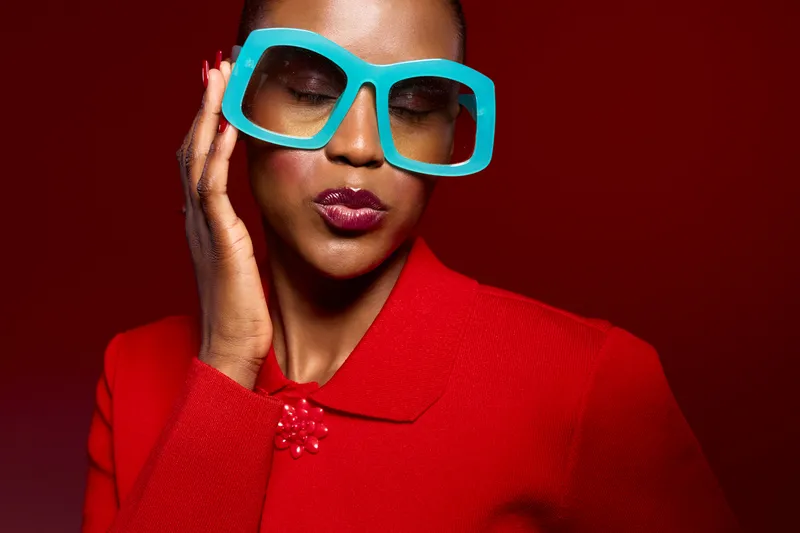 Stylish dark skinned African American woman wearing bright red coat and large turquoise sunglasses poses with closed eyes on red background. Fashion, beauty, and modern style concept.