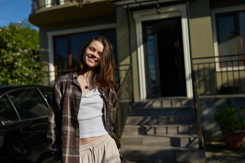 Cheerful woman outdoors, casual lifestyle moment, smiling with relaxed posture in a sunlit urban setting, light breeze, natural colors, building facade in background