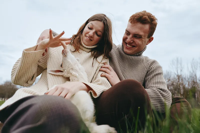 Smiling couple with goat kid in a grassy field, wearing knitted sweaters, enjoying a gentle moment. Soft natural light creates a warm, cozy mood focused on lifestyle, outdoors, affection,