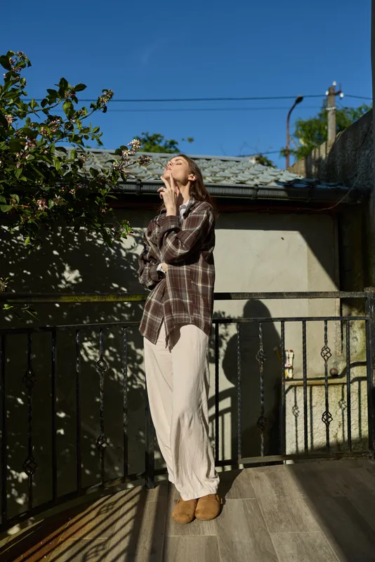 Cheerful woman on a sunny balcony, wearing a plaid shirt and light trousers, looking up with a gentle smile, relaxed pose, warm natural colors, soft shadows, casual lifestyle