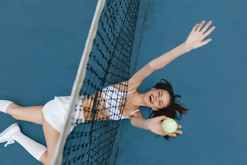 Energetic young woman playing tennis, smiling and reaching for a ball on a blue court in stylish athletic wear, embodying joy and sportiness