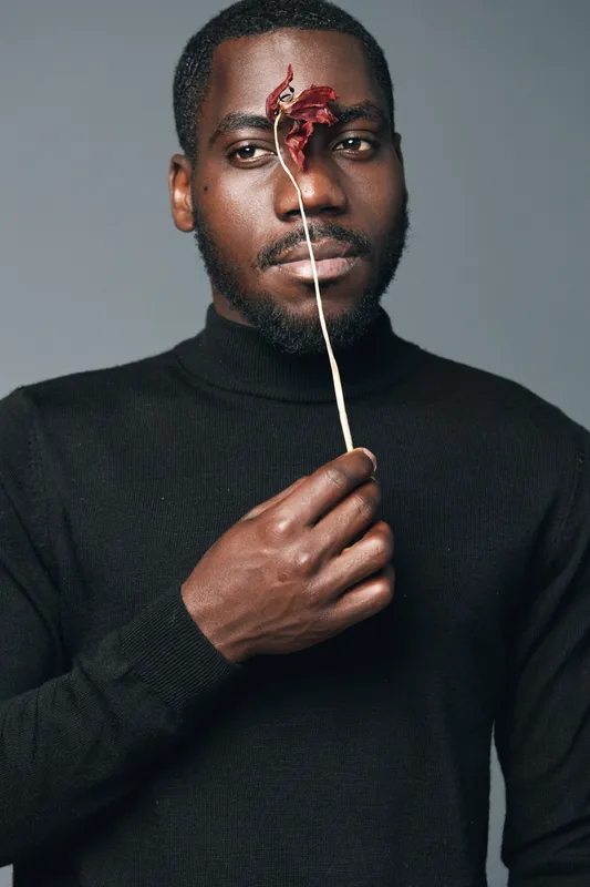 African man with dark skin wearing black sweater holding dried flower in front of face in creative conceptual visual style studio portrait with gray background.