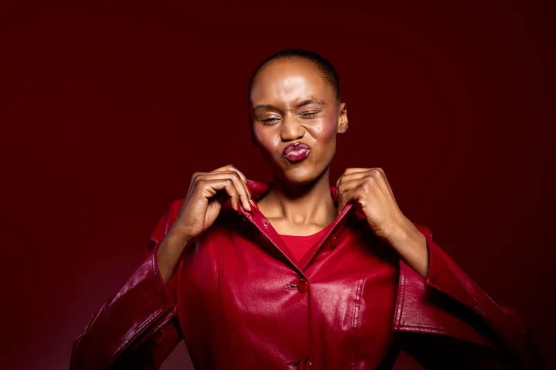 Confident dark skinned African woman in red leather jacket poses against red background, making playful face expression. Concept of fashion, attitude, and bold style for modern woman.
