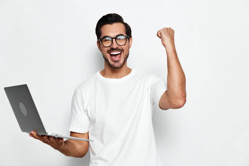 Happy smiling young man with glasses celebrates success while holding a laptop in his hand, isolated on a plain solid background. People lifestyle concept.