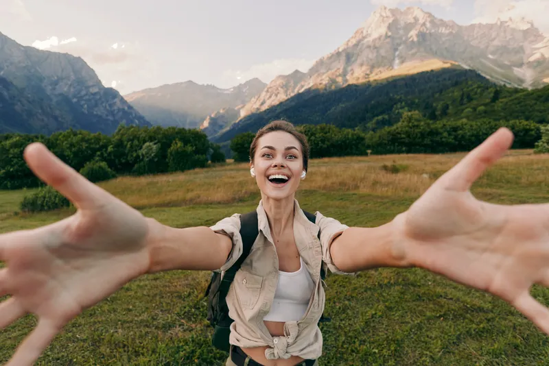 Joyful woman in an outdoor lifestyle scene, smiling and reaching toward the camera, wearing a backpack on a grassy meadow with mountains, conveying freedom and adventure in nature.