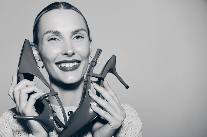 Smiling white woman with fair skin holding high heel shoes close to face in studio. Concept of fashion, style, and footwear.