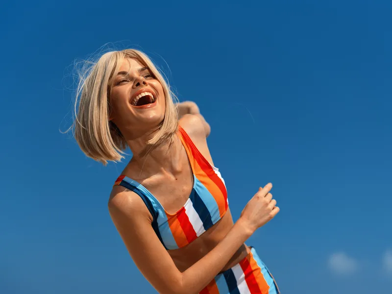 joyful young woman wearing colorful striped swimsuit laughing outdoors, enjoying bright sunny day, close-up portrait with clear blue sky background, summer fun and happiness