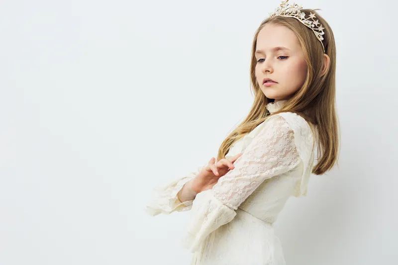 Elegant girl in a white dress with a crown, showcasing confidence and grace against a soft white background, perfect for themes of beauty and innocence