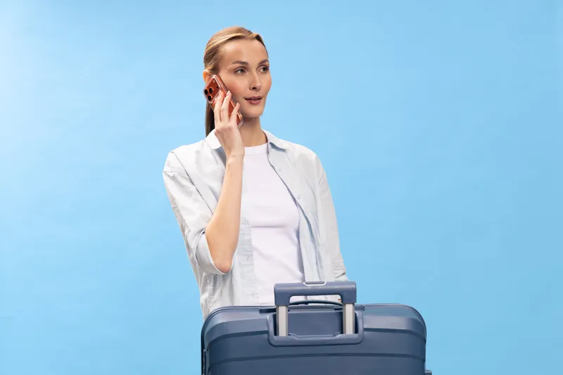 Young woman in casual clothes holds a suitcase and talks on a smartphone. Blue background, travel and communication concept with relaxed female traveler preparing for a trip.