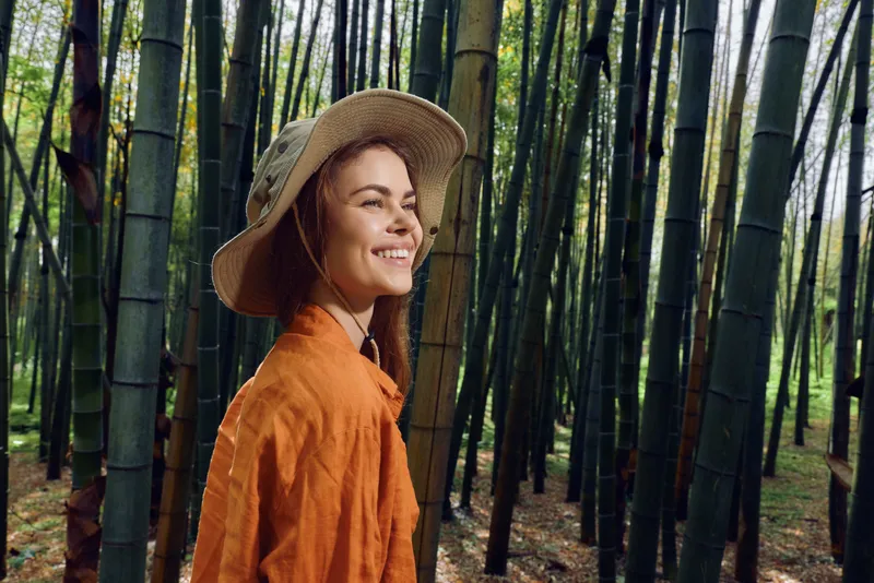 Woman bamboo forest smile hat portrait, young woman in orange shirt and straw hat walking through dense bamboo grove, smiling and looking sideways during outdoor nature excursion.