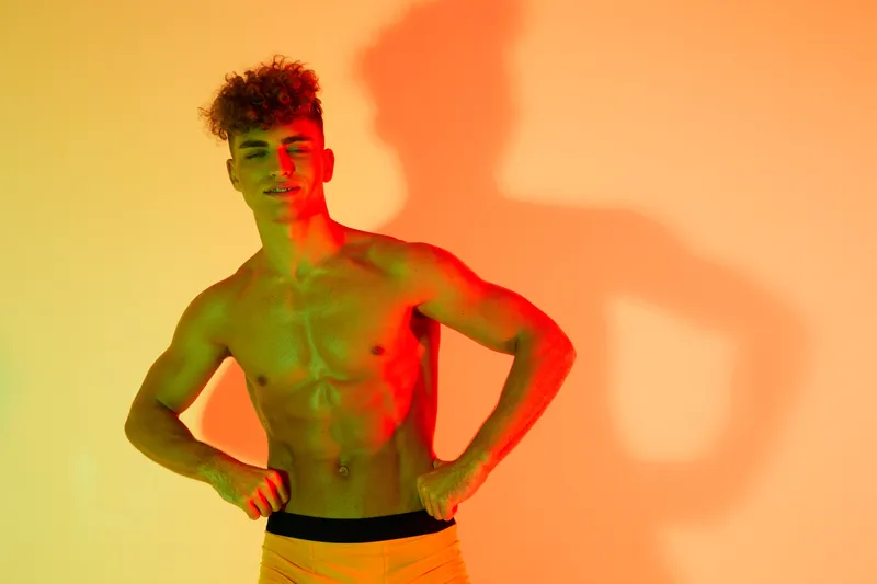 Young muscular man posing shirtless with hands on hips under colorful lighting and shadow on wall, fitness and body confidence concept with vibrant orange and green tones.