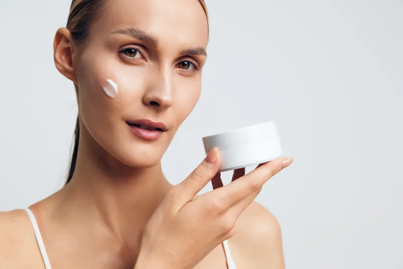 Young woman applying white face cream on cheek holding a cosmetic jar. Skincare routine concept with natural beauty and clean skin on a plain light background. Soft natural lighting.