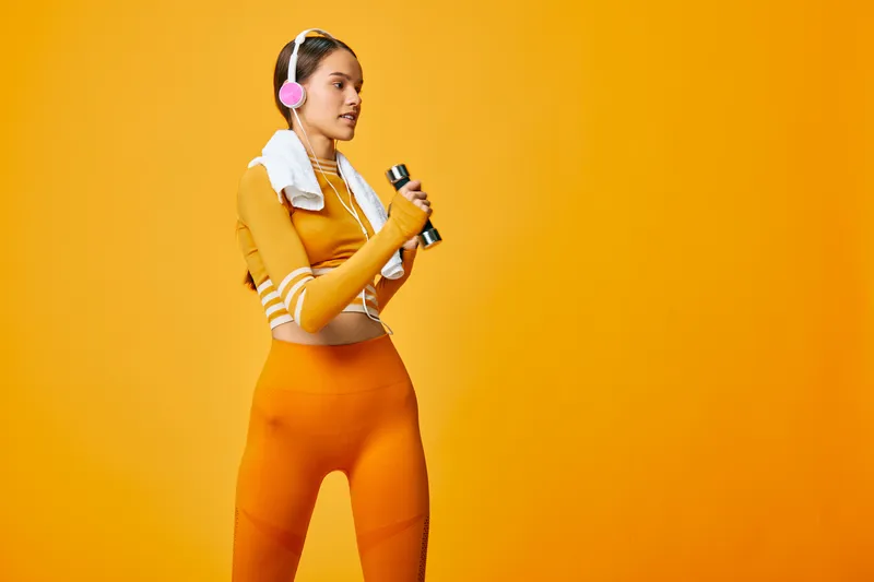 Fitness woman in orange athletic wear with a towel and headphones holds a dumbbell against a vibrant orange studio backdrop, ready for a workout, conveying energy, focus and healthy sport lifestyle.