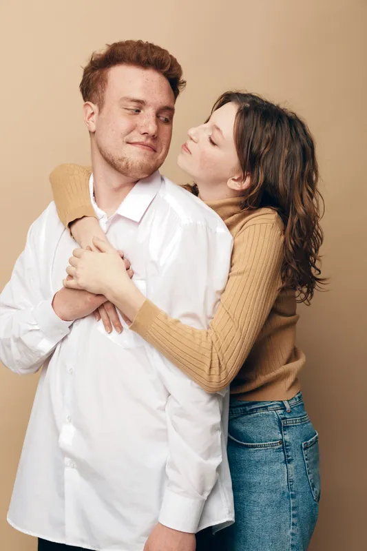Young couple in casual clothes embraces tenderly against beige background. Woman hugs man from behind showing affection and warmth. Concept of love, relationship, and intimacy.