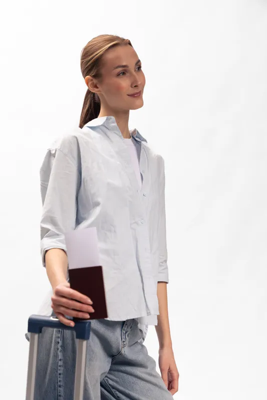 Young woman with light brown hair holds passport and suitcase handle standing on white background. Concept of travel, tourism, and vacation preparation for a trip.