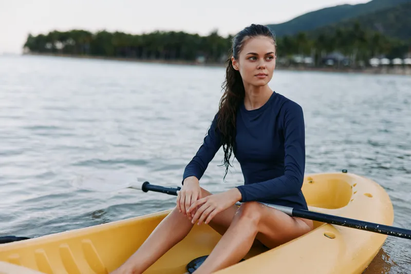 Serene portrait of a lone woman in a navy top seated in a yellow rowing boat, hands resting on the oar, calm water ahead, distant shore behind, timeless outdoor scene.