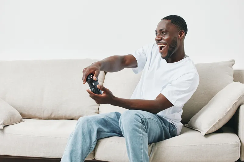 Happy young Black man playing video games on a couch, wearing a white t shirt and jeans, expressing joy and excitement in a modern living room