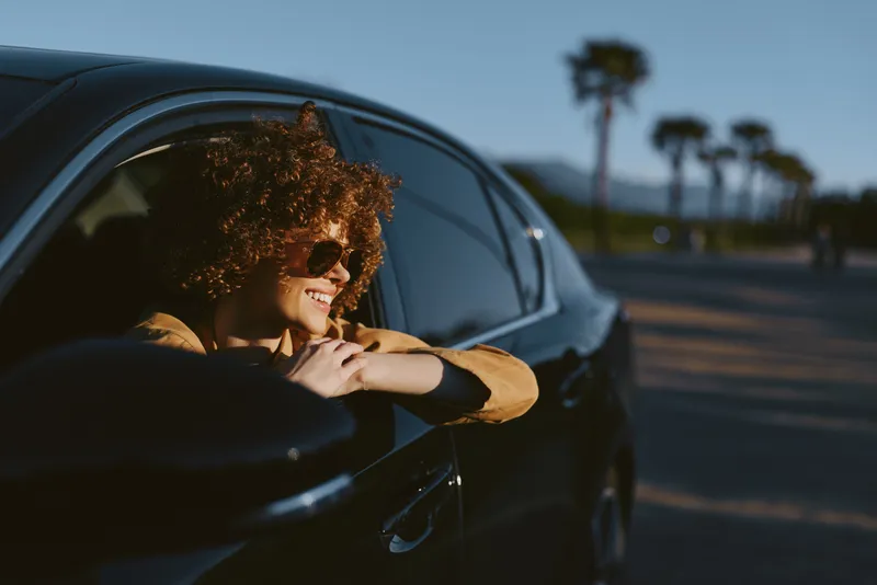 Cheerful woman leaning from a car window, enjoying a sunny outdoor moment, retro film color, relaxed pose, breezy hair, warm hues, palm trees in the distance, casual lifestyle scene