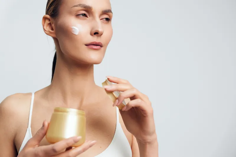 A young woman applies moisturizing cream to her face using a small spatula. She holds a gold jar of skincare product, demonstrating a beauty routine in a bright studio setting.