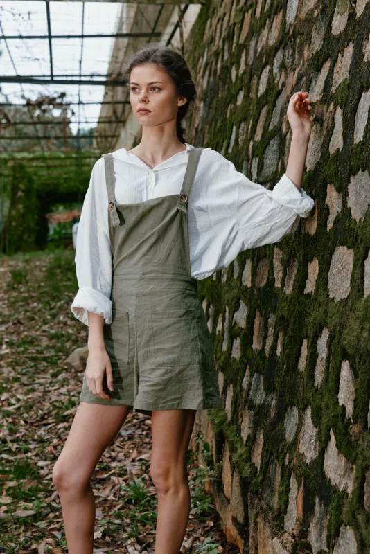 Young woman in an olive green dress and white blouse standing against a stone wall, conveying a natural, serene feeling in a lush outdoor setting