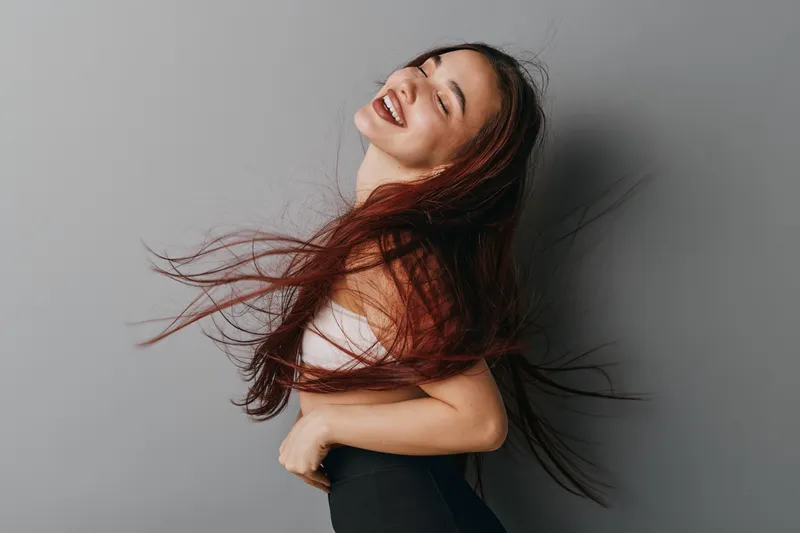 Woman with long hair smiling and turning with motion against gray background. Joyful female model in casual clothes with natural expression and flowing hair in studio.