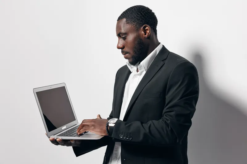 Black man with dark skin wearing black suit and white shirt using laptop computer for work or communication on white background. Business professional, technology and digital device.
