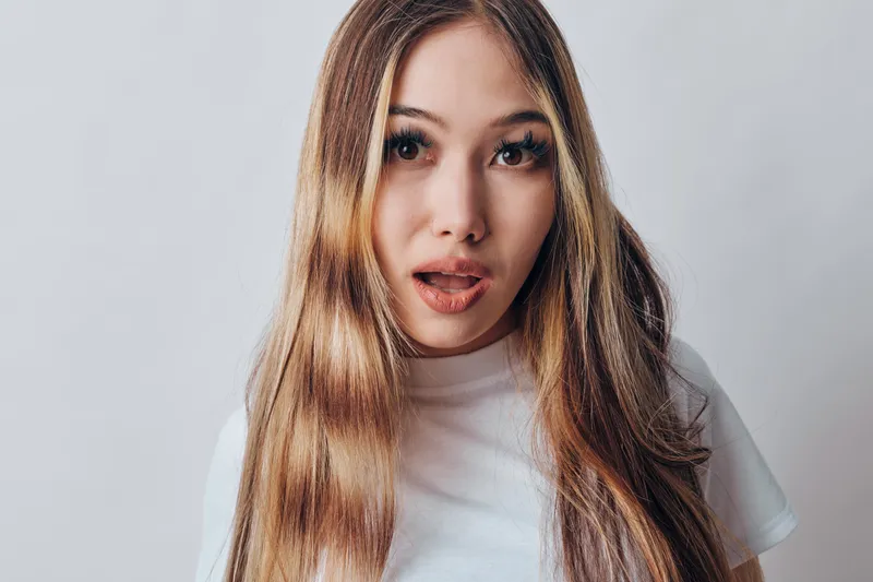 Young woman with long brown hair and white shirt poses with surprised expression against simple light background indoors. Closeup portrait capturing natural beauty and emotion.