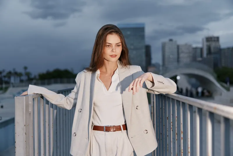 Confident woman in light business suit outdoors with urban skyline background during overcast day, modern fashion, professional style, city atmosphere, casual, elegant look