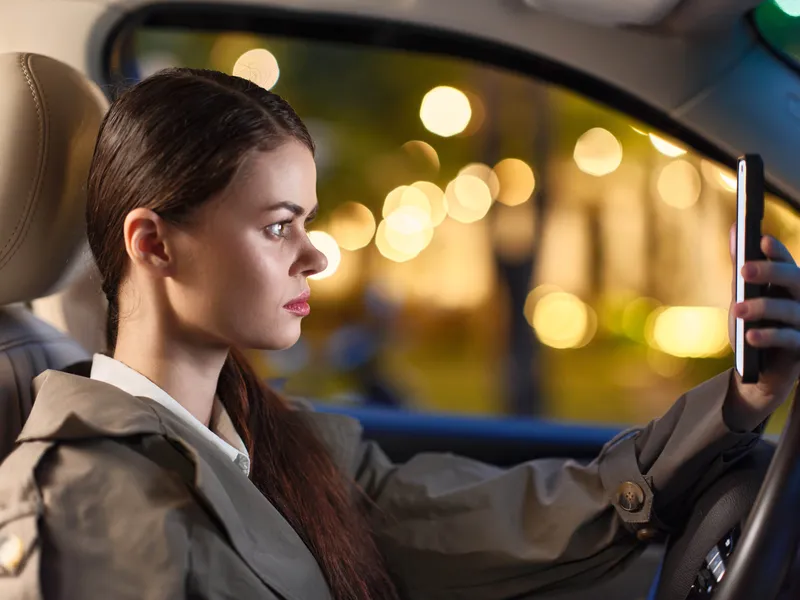 Женщина в автомобиле ночью пользуется телефоном: она сидит на водительском сиденье машины в темное время суток, сосредоточенно глядя на экран смартфона, который освещает ее лицо в темноте
