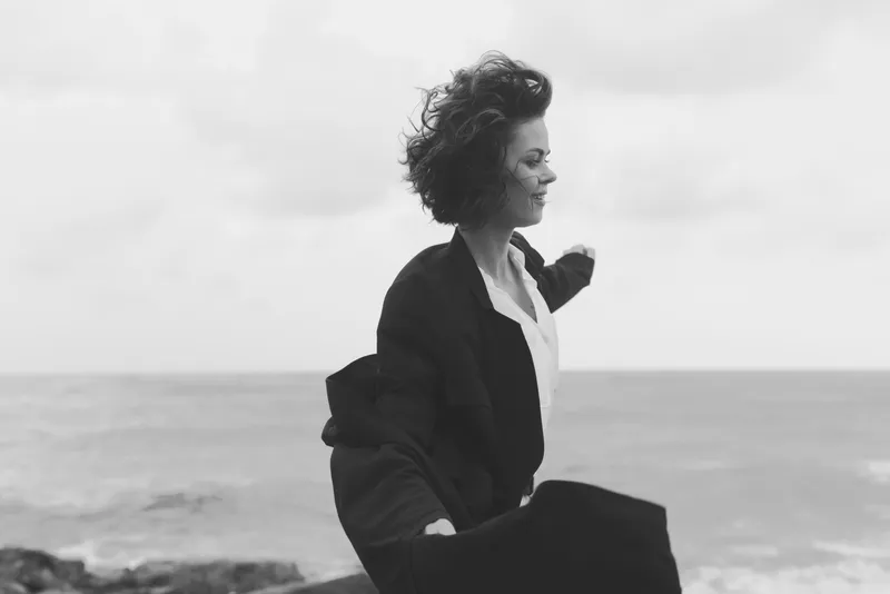 A woman stands on a windy coastline with a flowing coat, sea in the background, black and white mood, motion and vast sky