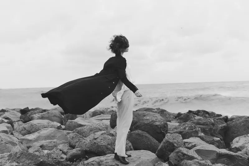 Woman wearing a flowing cape and coat stands on rugged rocks by the sea, wind sweeping garments, monochrome outdoor scene with dramatic mood and fashion forward silhouette.