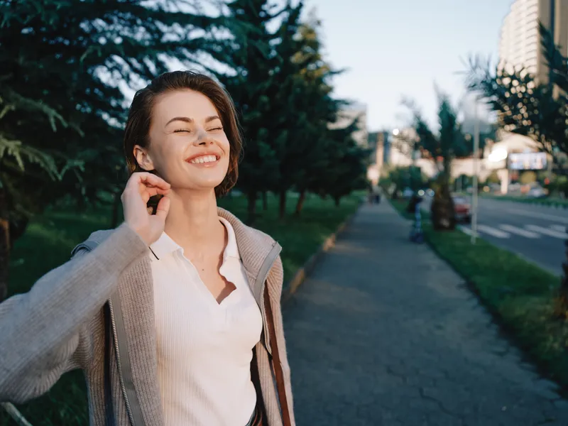 A cheerful woman is outdoors along a sunny city sidewalk in casual attire, smiling with a relaxed pose, radiating confidence and positive energy in a modern urban environment.