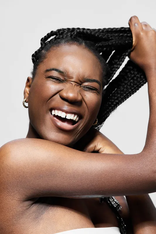 Smiling African woman with braided hair enjoying self care, showcasing beauty and positivity in a studio setting with neutral background
