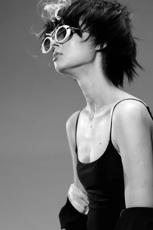 Black and white portrait of a stylish woman wearing round sunglasses exhaling smoke in edgy fashion style.