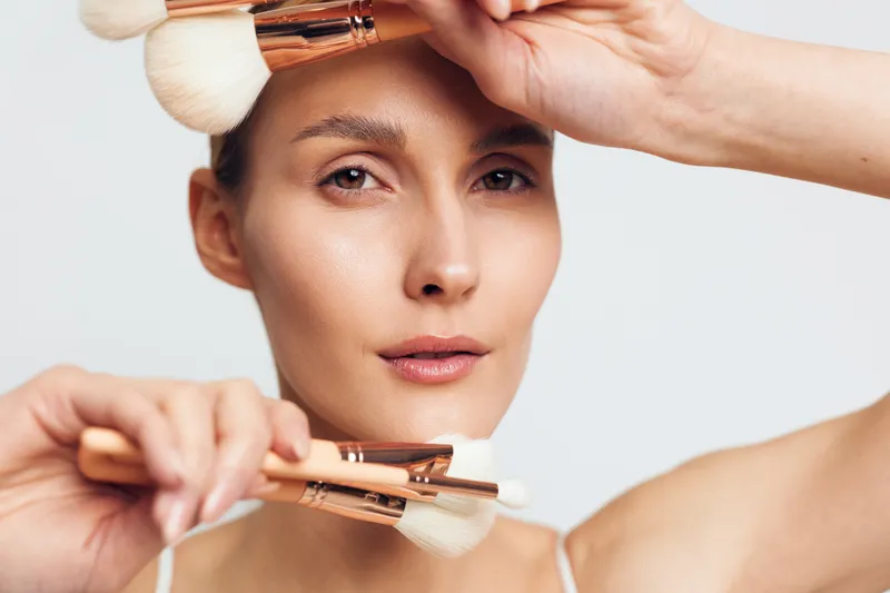 Young woman applying makeup with soft brushes, closeup portrait, natural skin tone, focused expression, clean background, beauty routine concept, gentle lighting