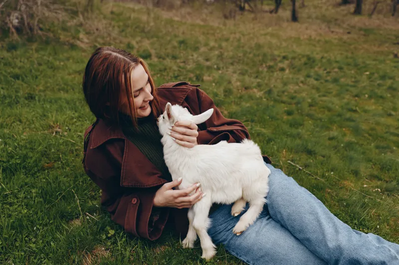A woman sits on grass, hugging a young goat kid in a brown jacket, smiling softly. A cozy lifestyle moment highlighting bonding, care, and gentle companionship with animals outdoors.