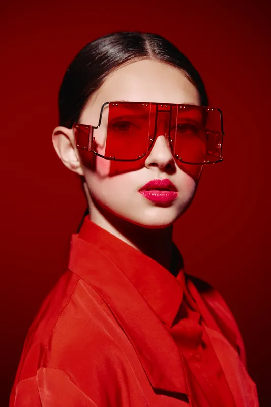Young woman wearing oversized red sunglasses poses confidently against a vivid red backdrop. Fashion concept highlighting bold beauty and creative styling.