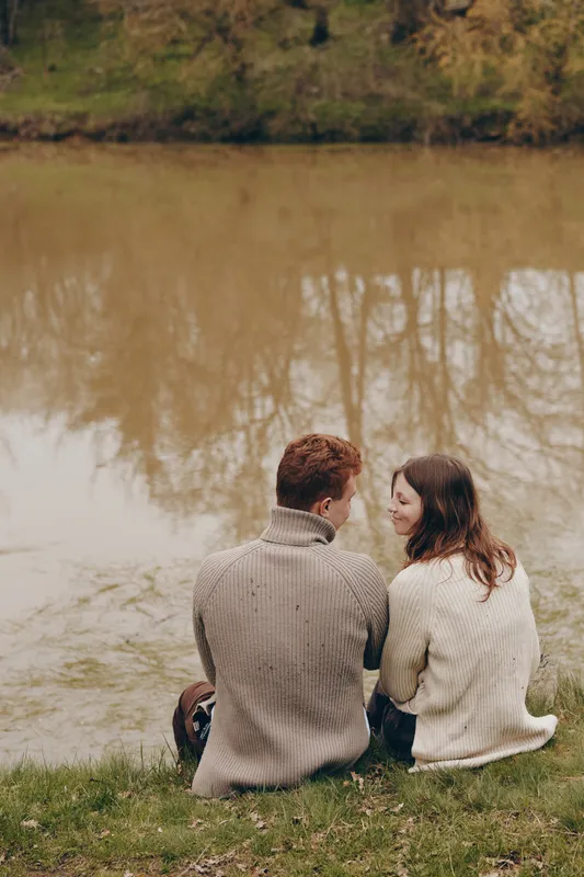 A couple sits on the grassy riverbank, wearing sweaters, facing each other with smiles, enjoying quiet companionship and parklike scenery near calm water, a candid moment of togetherness in soft