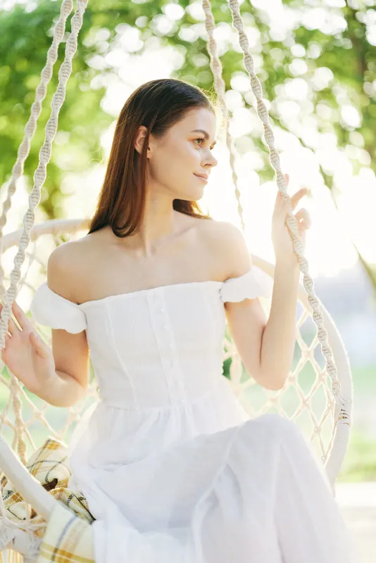 Young woman in a white off shoulder dress sitting on a swing in a sunny park, showcasing a serene expression amidst nature s greenery