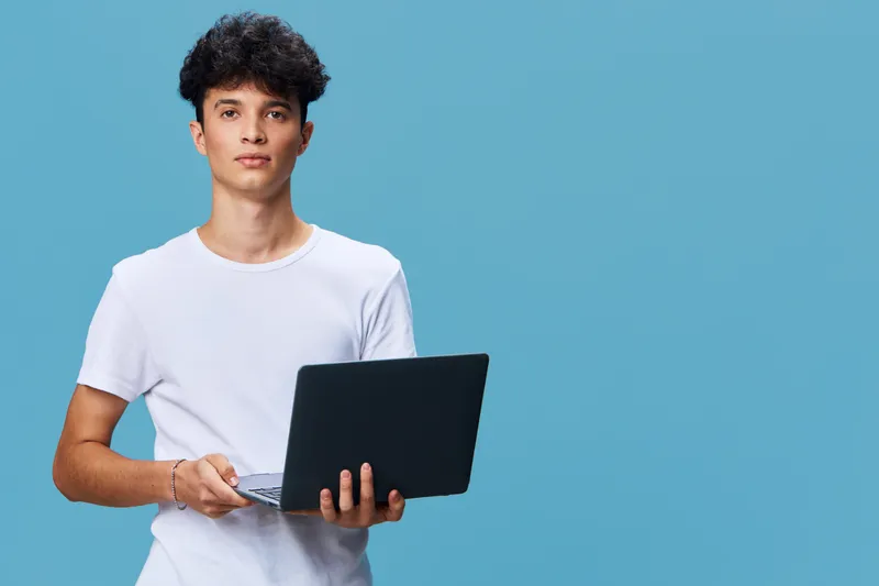 young man holding laptop, casual white t-shirt, standing against blue background, focused expression, technology and work concept
