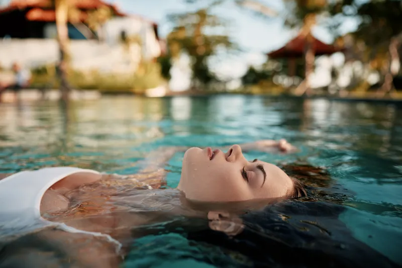 Young woman in white swimsuit floating on water surface with closed eyes, relaxing in outdoor pool. Concept of lifestyle, wellness, and peaceful leisure under natural light.