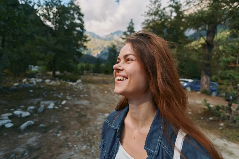 A cheerful woman stands outdoors in a natural setting, smiling brightly as she gazes upward. Dressed in a denim jacket, she exudes relaxed confidence and optimism in daylight.