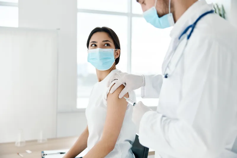 Врач в медицинском халате делает укол вакцины в плечо пациента в медицинской маске