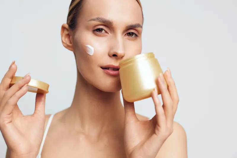 Young woman with blonde hair applies moisturizing cream on her cheek while holding a golden jar and lid in a bright studio setting with a neutral background.