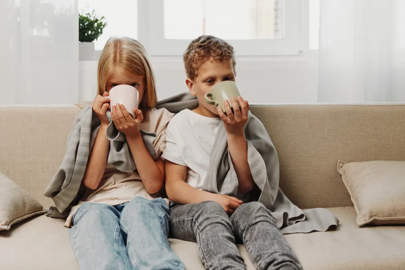 Two children wrapped in blankets sit on a sofa sipping hot drinks, cozy atmosphere with natural light from window. Concept for warmth, comfort, and family moments.