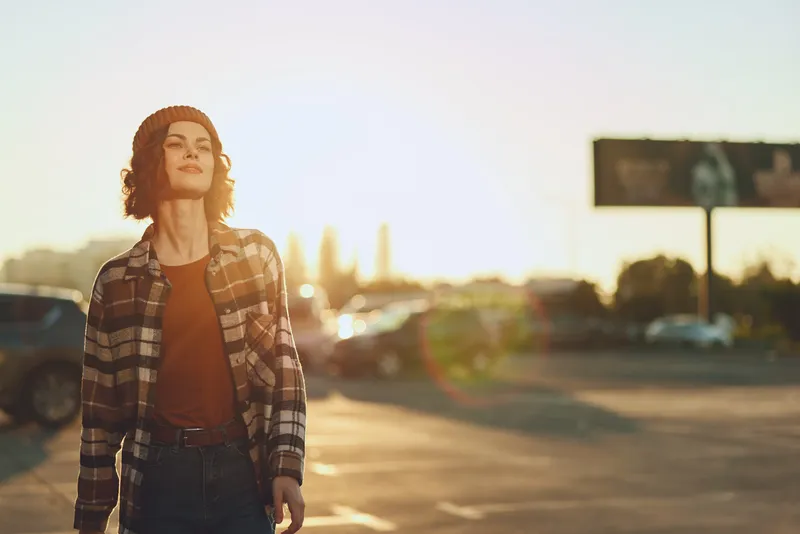 Woman walking in urban parking lot at golden hour with sunlight flare, candid lifestyle portrait of authenticity and mindful living, beanie and casual plaid outfit, relaxed mood.