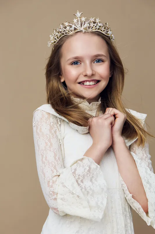 A joyful young girl in a white lace dress and a sparkling crown smiles warmly, showcasing happiness and elegance against a soft beige background