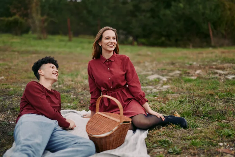 A young couple enjoys a relaxed picnic on a blanket in a grassy field, sharing warm smiles and quiet conversation. The scene conveys genuine connection, authenticity and credibility in everyday