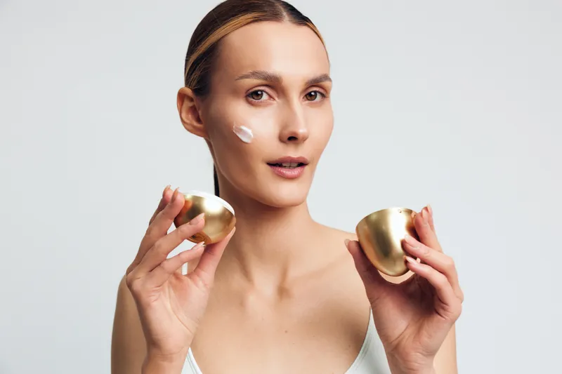 Young woman studio portrait, skincare beauty routine on face, holding cosmetics jar, serum jar and moisturizer jar, fresh glowing complexion, minimal white background, calm confident mood